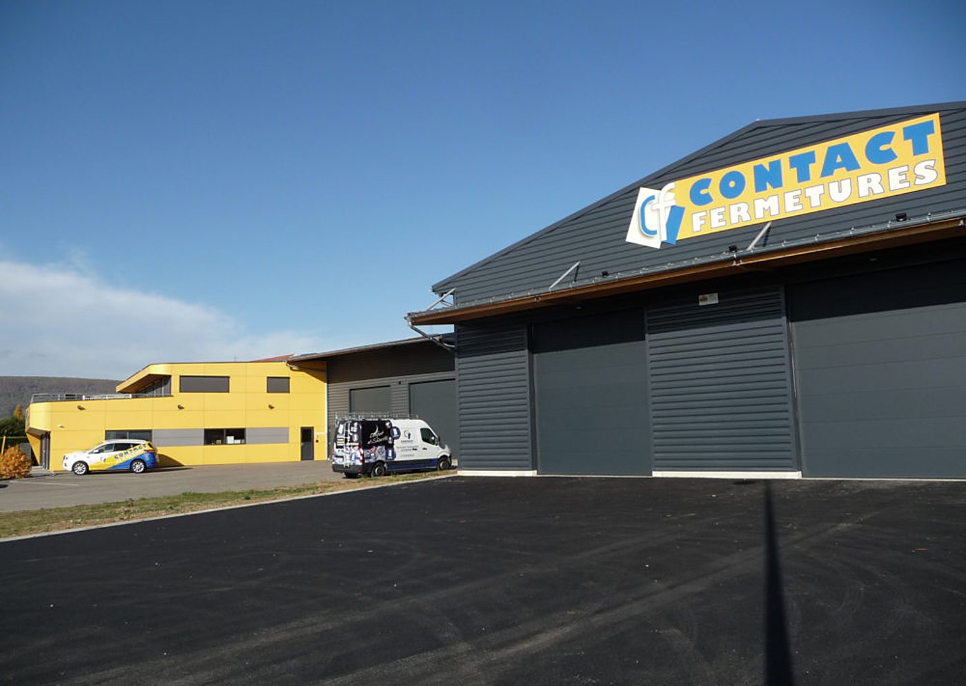 Façade d'un bâtiment industriel gris portant l'enseigne Contact Fermetures, à côté d'un bâtiment jaune, avec une camionnette et une voiture garées sur un parking fraîchement goudronné.
