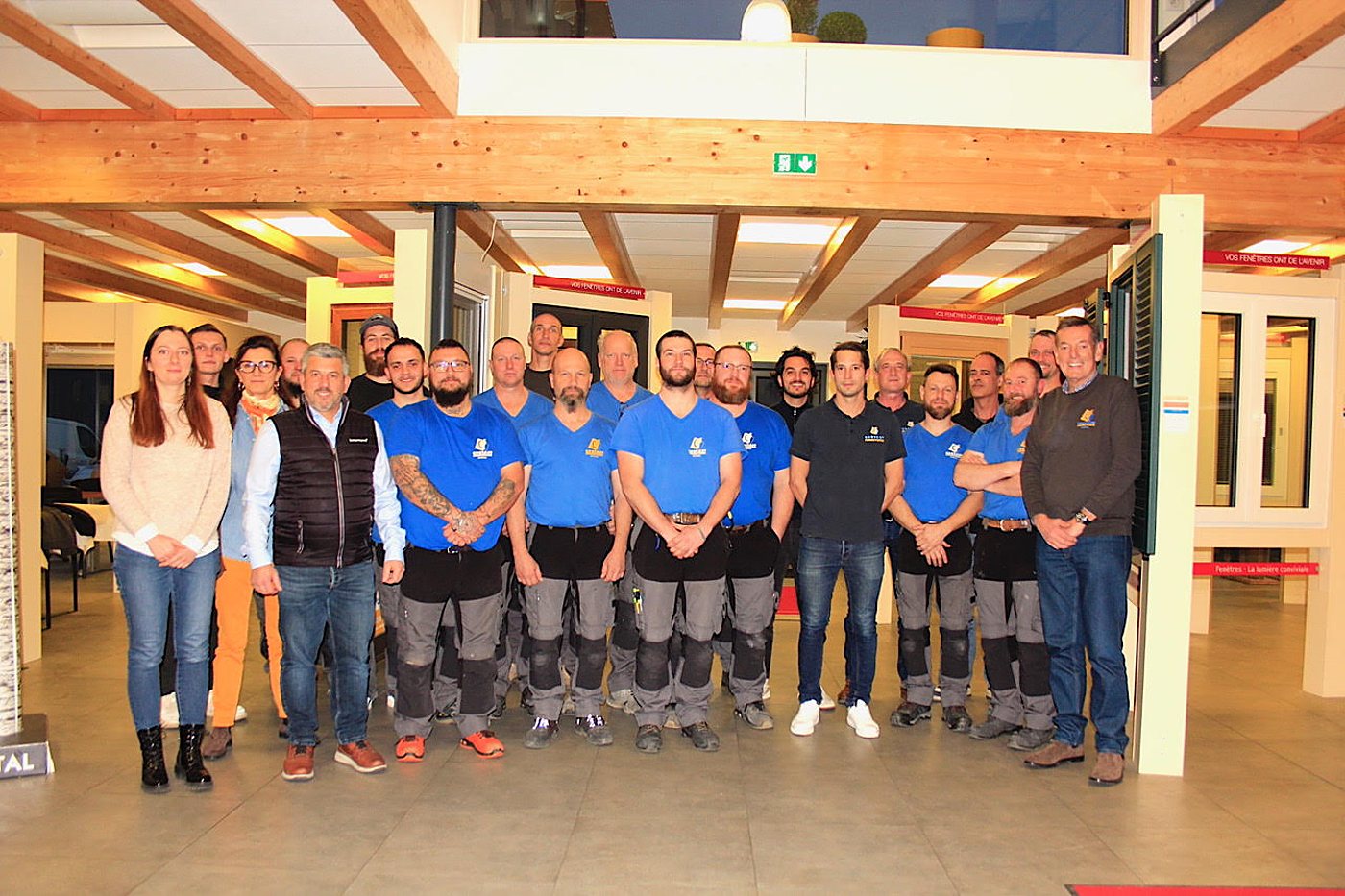 Une vingtaine de personnes, dont beaucoup portent un t-shirt bleu, posent ensemble dans un showroom de fenêtres aux poutres en bois.