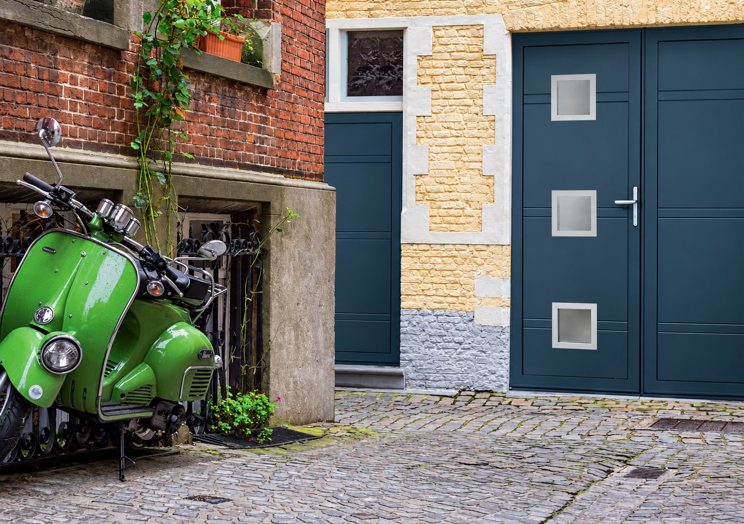 Un scooter vert vintage est garé dans une cour pavée, près d'une porte de garage grise à petites fenêtres carrées et d'un mur de briques.
